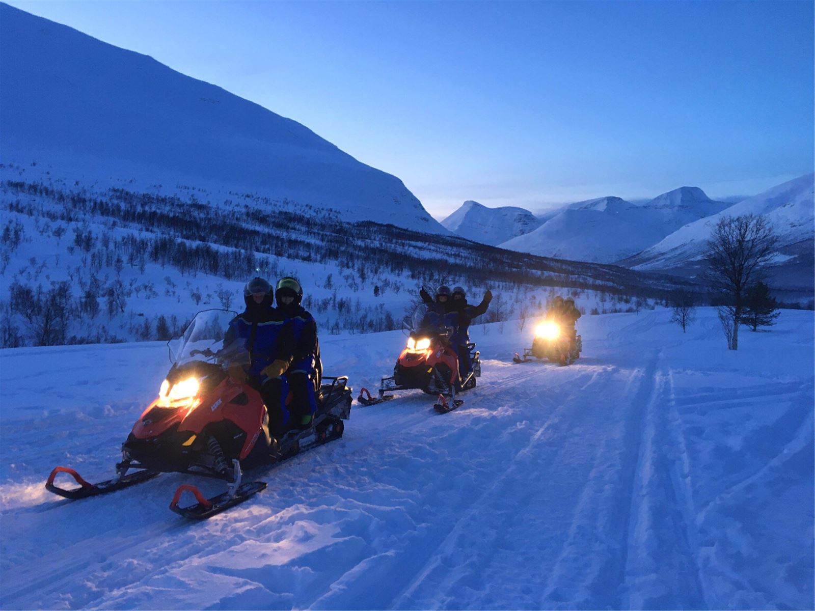 Three snowmobiles driving through snow in blue twilight