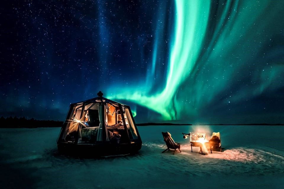 AuroraHut glass igloo with a campfire under green northern lights